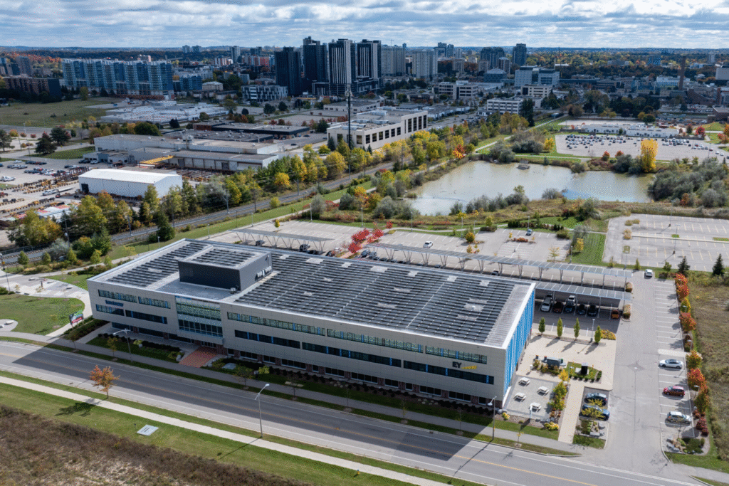Aerial view of evolv1 building in Waterloo showcasing its sustainable design, rooftop solar panels, spacious parking lot, and surrounding green spaces with city skyline in the background.