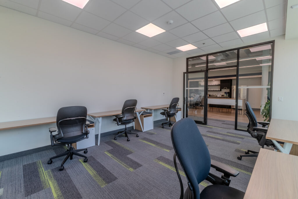 Premium 5-person office suite at Cora GRID coworking space featuring ergonomic desks and chairs, floor-to-ceiling windows with natural light, scenic autumn views, and modern interior design with glass-walled meeting room and patterned carpet.