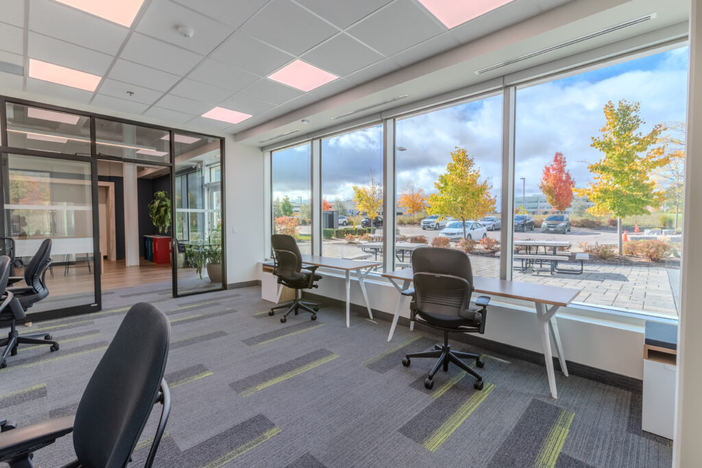 Premium 6-person office suite at Cora GRID coworking space featuring ergonomic desks and chairs, floor-to-ceiling windows with natural light, scenic autumn views, and modern interior design with glass-walled meeting room and patterned carpet.